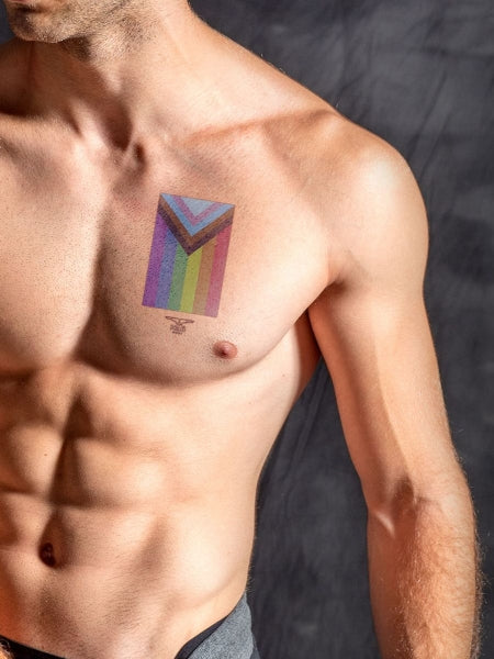ansicht einer glatten muskolösen männerbrust vor einer dunklen wand der ein temporäres tattoo von mister b auf der linken brust und der pride LGBTQ+ flagge in regenbogen auf dem gay körper