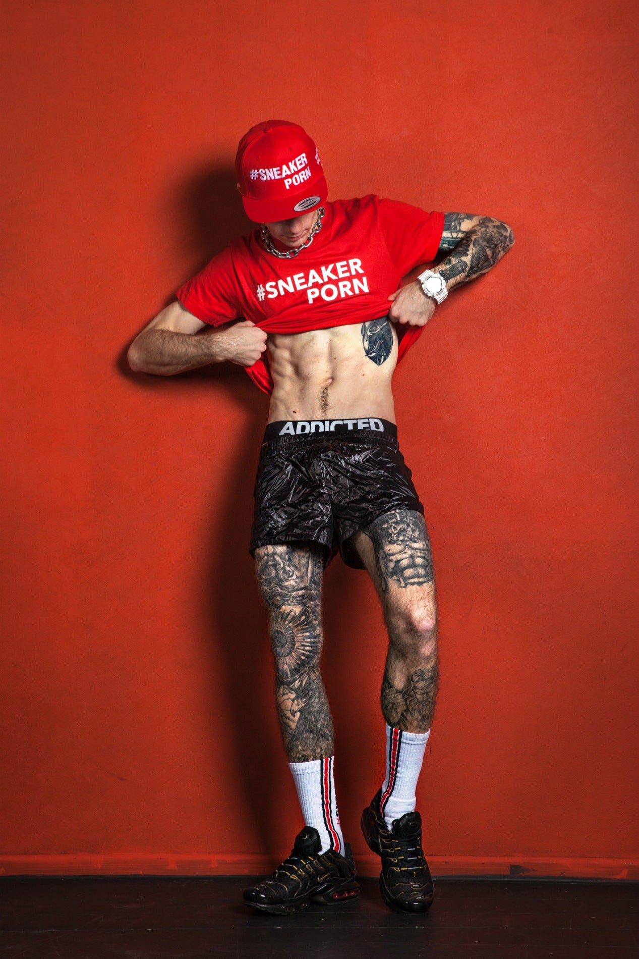 sexy boy mit addicted jock und sneakerporn socken von sk8erboy trägt ein rotes #sneakerporn t-shirt und basecap vor einer roten wand