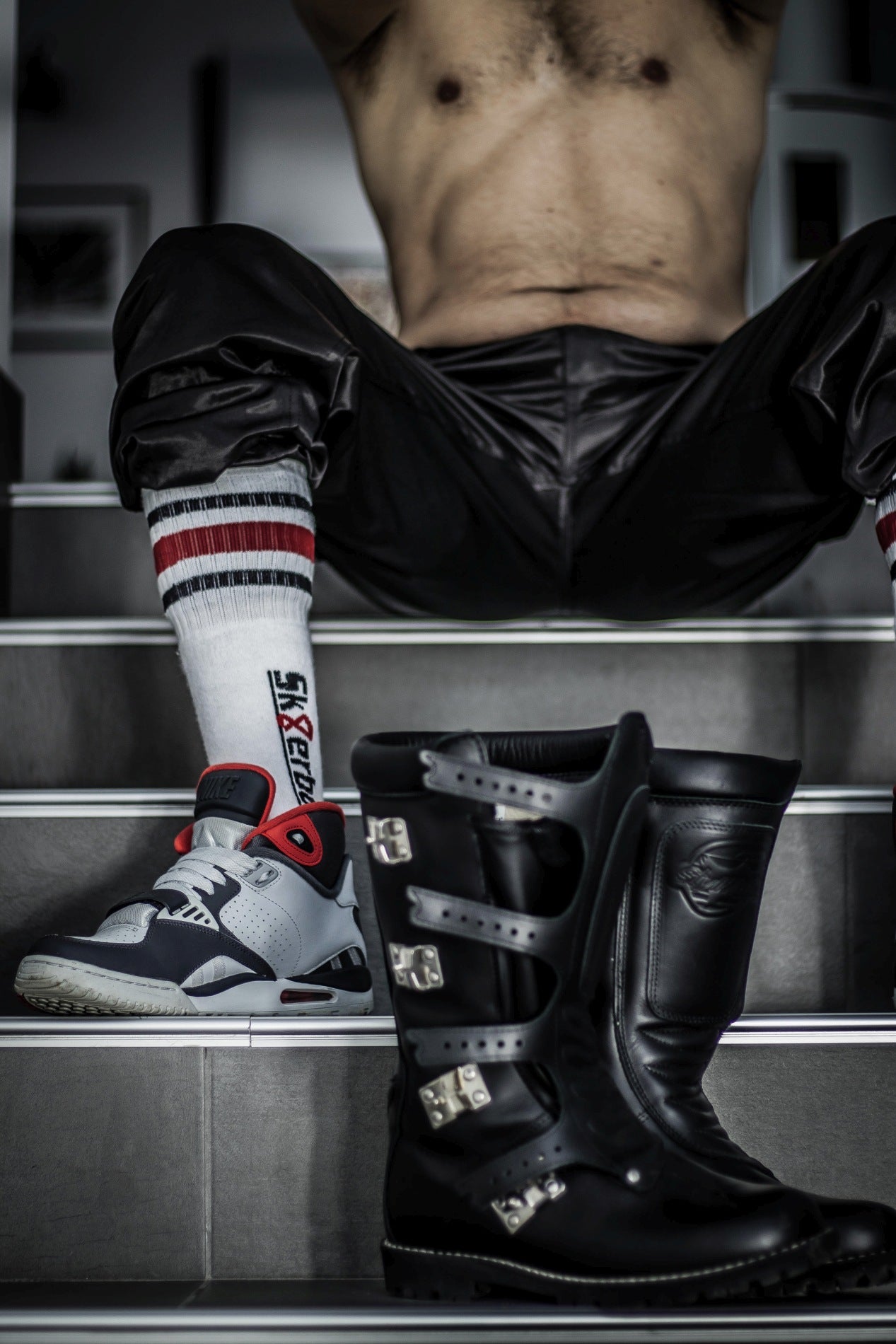 sportlicher gay sitzt auf einer treppe und traegt in seinen sneaker sk8erboy tube socks in schwarz weiss rot mit grossem logo an der seite und biker leder boots vor sich