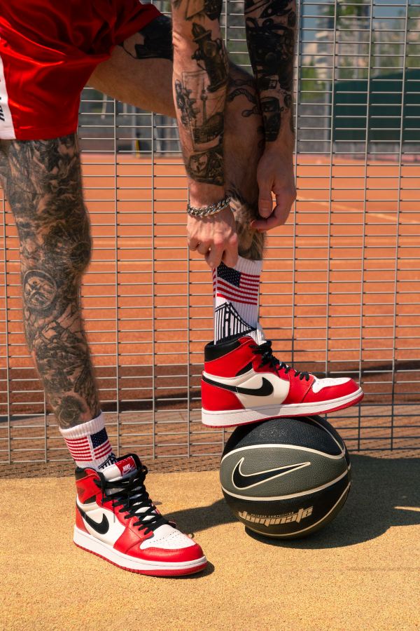 junger sportlicher gay in nahaufnahme mit tattoos an beinen und armen zieht sich die weissen socken mit usa flagge hoch in seinen roten nike turnschuhen auf einem basketball feld