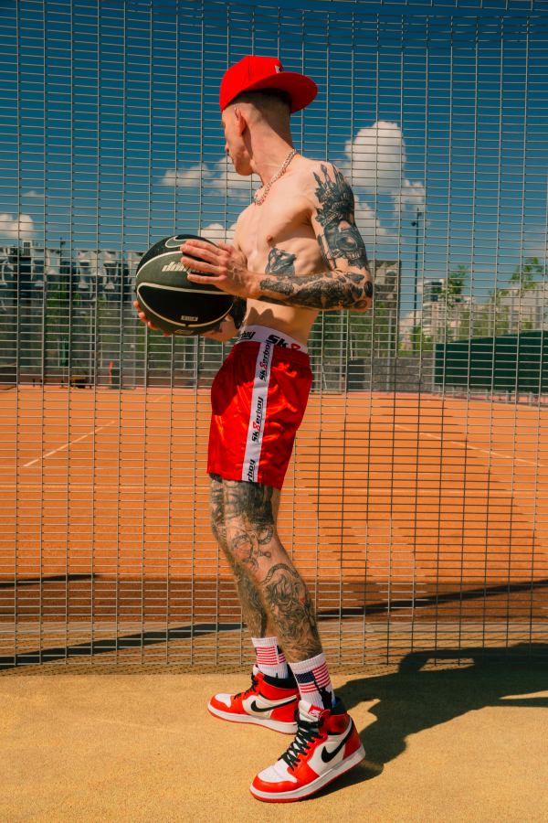 junger sportlicher gay in nahaufnahme mit tattoos an beinen und armen mit usa flagge auf seinen socken in seinen roten nike turnschuhen auf einem basketball feld haelt einen basketball in den haenden