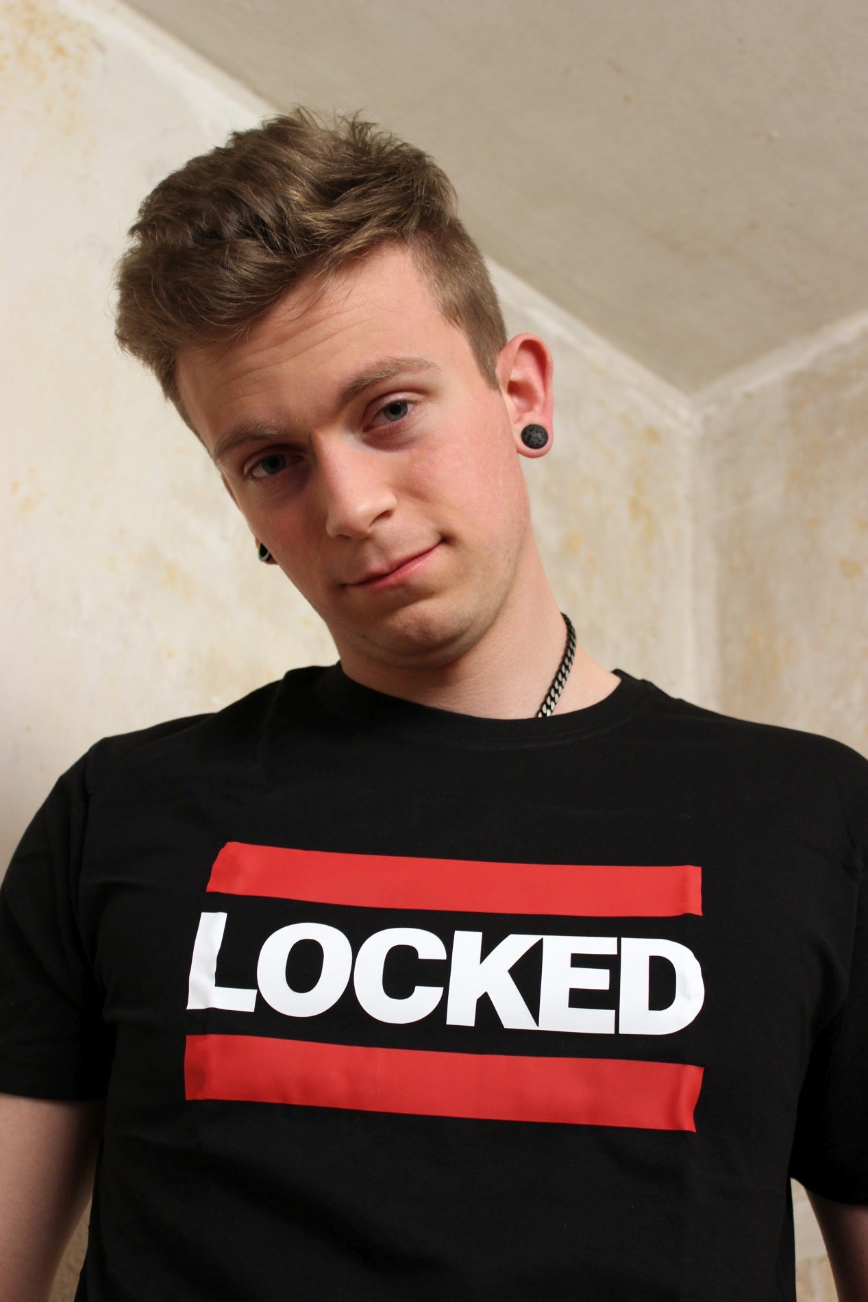 junger gay traegt schwarzes sk8erboy t-shirt mit großen weissen buchstaben LOCKED auf der vorderseite mit roten balken darueber und darunter vor einer weissen wand fuer gay und fetisch